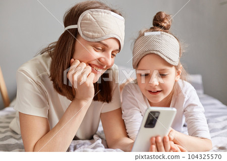 Cheerful woman and her daughter in sleeping mask laying on bed and using mobile phone looking at device screen with smile enjoying checking social networks. Cheerful woman and her daughter in sleeping mask laying on bed and using mobile phone looking at device screen with smile enjoying checking social networks. 104325570