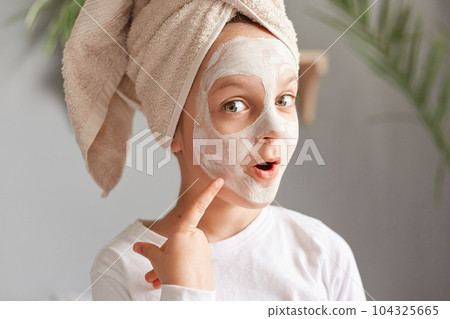 Astonished little girl with towel on head with cosmetic mask pointing at her face with excited look doing skin care procedures wearing white shirt. Astonished little girl with towel on head with cosmetic mask pointing at her face with excited look doing skin care procedures wearing white shirt. 104325665