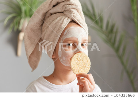 Funny cute charming little girl with towel on head with cosmetic mask holding covering mouth with cosmetic sponge looking at camera. Funny cute charming little girl with towel on head with cosmetic mask holding covering mouth with cosmetic sponge looking at camera. 104325666