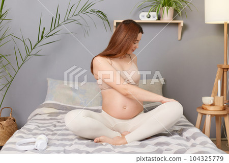 Attractive pregnant brown haired caucasian woman sitting in bed resting doing stretching exercises for her body taking care of her health. Attractive pregnant brown haired caucasian woman sitting in bed resting doing stretching exercises for her body taking care of her health. 104325779
