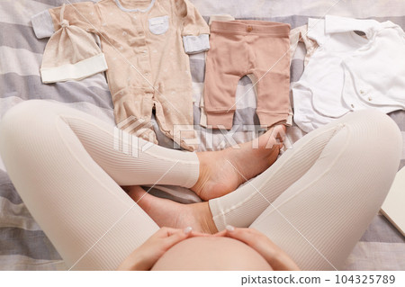 Unknown unrecognizable pregnant woman wearing leggins sitting in bed among tiny baby's clothing holding her bare belly packing things. 104325789