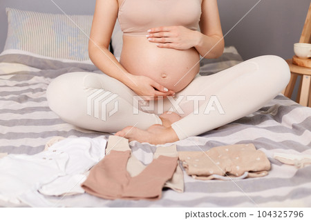 Unknown pregnant woman wearing beige top and leggins sitting in bed surrounded with tiny kid's clothing preparing for giving birth. 104325796