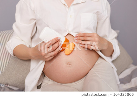 Unrecognizable pregnant woman wearing white shirt sitting in bed using cream for stretch prevention talking care of her skin during pregnancy. 104325806
