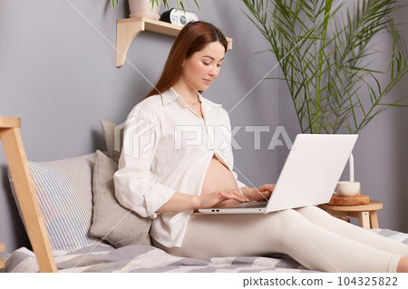Side view portrait of pregnant brown haired caucasian woman sitting in bed and working on laptop typing on keyboard answering e mails communicating online in social networks. 104325822