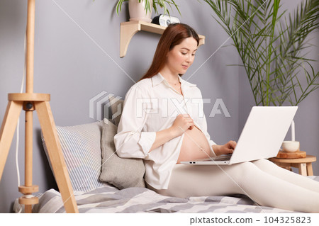 Pregnant woman with bare belly sitting on bed using laptop future mom expecting child working on computer answering email messages browsing social network. 104325823