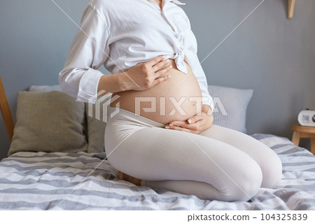 Healthy pregnancy. Expecting childbirth. Unrecognizable unknown pregnant woman wearing white clothing sitting in bed in light bedroom holding her bare belly. Healthy pregnancy. Expecting childbirth. Unrecognizable unknown pregnant woman wearing white clothing sitting in bed in light bedroom holding her bare belly. 104325839