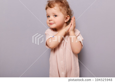 Indoor shot of cute curios little toddler caucasian girl posing isolated over gray background, baby with blond curly hair keeps palms together, free space for advertisement. Indoor shot of cute curios little toddler caucasian girl posing isolated over gray background, baby with blond curly hair keeps palms together, free space for advertisement. 104325948