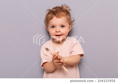 Charming little girl holding toothbrush isolated on gray background looking at camera holding toothbrush trying to brush teeth by herself. 104325958