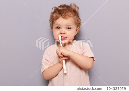 Funny little child baby brushing teeth isolated over gray background oral care using kid's toothbrush looking at camera, wearing t-shirt. 104325959