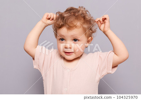 Cute blonde curly haired toddler baby little caucasian girl posing isolated over gray background touching her wavy hair looking away at something interesting. 104325970