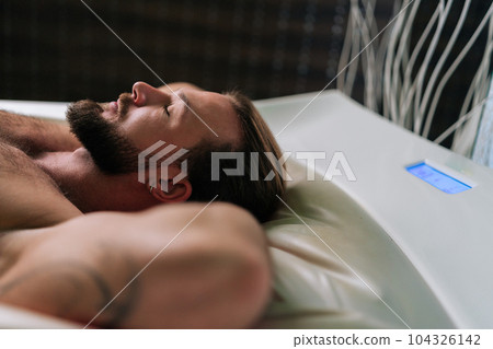 Closeup side view face of relaxed young man with earring in ear lying relaxing on electric massage bed with closed eyes, enjoying spa treatment at spa salon. Muscular male having rest enjoying weekend Closeup side view face of relaxed young man with earring in ear lying relaxing on electric massage bed with closed eyes, enjoying spa treatment at spa salon. Muscular male having rest enjoying weekend 104326142