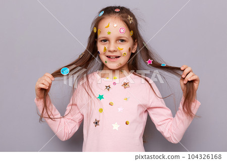 Beautiful cute little girl covered colorful kid's stickers standing isolated over gray background pulling her hair aside looking at camera with smile having fun expressing positive emotions. 104326168