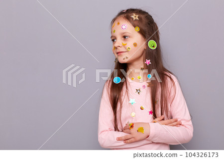 Optmistic smiling little girl covered colorful kid's stickers standing isolated over gray background lookign away and advertisement area, copy space for promotion. 104326173