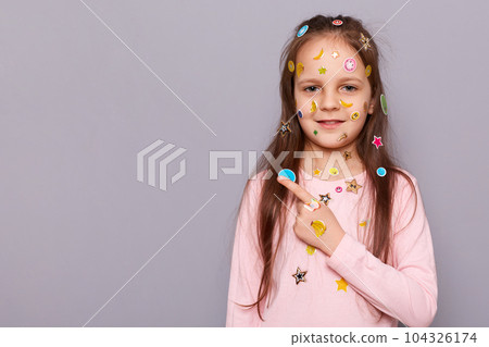 Cute charming adorable little girl covered colorful kid's stickers standing pointing away ant free space for advertisement posing isolated over gray background. Cute charming adorable little girl covered colorful kid's stickers standing pointing away ant free space for advertisement posing isolated over gray background. 104326174