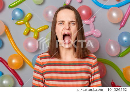 Angry despair woman wearing striped dress standing against gray wall with balloons decoration screaming with hate and aggression looking at camera open mouth. Angry despair woman wearing striped dress standing against gray wall with balloons decoration screaming with hate and aggression looking at camera open mouth. 104326262