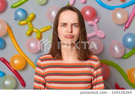 Sad displeased woman wearing striped dress standing against gray wall with balloons decoration looking at camera with pout lips disappointed facial expression. Sad displeased woman wearing striped dress standing against gray wall with balloons decoration looking at camera with pout lips disappointed facial expression. 104326264