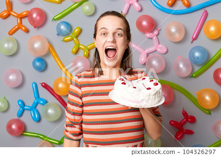 Happy joyful cheerful woman with brown hair wearing striped dress holding cake standing against gray wall decorated with colorful balloons posing with sweet dessert on her face, screaming with joy. Happy joyful cheerful woman with brown hair wearing striped dress holding cake standing against gray wall decorated with colorful balloons posing with sweet dessert on her face, screaming with joy. 104326297
