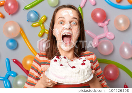 Amazed excited happy woman in striped dress standing against gray wall decorated with colorful balloons showing her cake celebrating birthday screaming excitement. 104326308