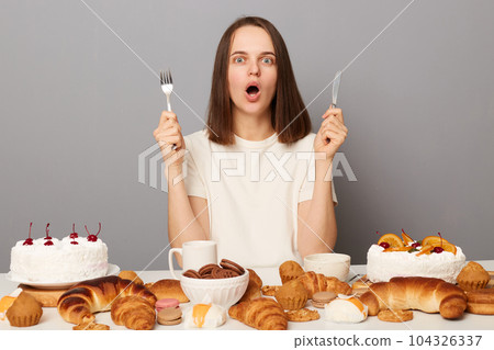 Shocked woman wearing white T-shirt sitting at table isolated over gray background eating junk food bakery looking at camera with big eyes and open mouth. 104326337