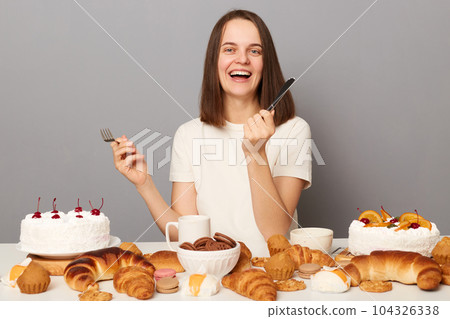 Extremely happy positive woman wearing white T-shirt isolated over gray background sitting at festive table among bakery sugary desserts holding fork and knife. 104326338