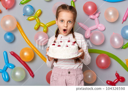 Amazed surprised cute little girl  with pigtails holding dessert cake in hands looking at camera with big eyes standing against gray wall decorated with colorful balloons. 104326598