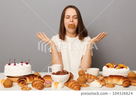 Funny woman wearing white T-shirt isolated over gray background sitting with cookies in mouth shrugging shoulders being hungry breaking diet. Funny woman wearing white T-shirt isolated over gray background sitting with cookies in mouth shrugging shoulders being hungry breaking diet. 104326624