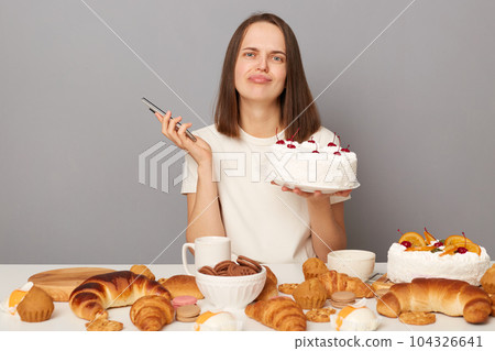 Dissatisfied sad woman wearing white T-shirt isolated over gray background sitting at festive table among pastry holding cake and smartphone in hands, looking at camera with pout lips. 104326641