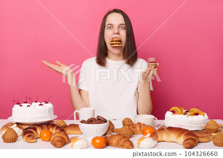 Funny hungry brown haired woman in white t shirt sitting at table with sweets isolated over pink background posing with mouth full of cookies. 104326660