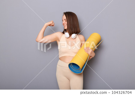 Strong young attractive caucasian smiling woman with yoga mat in her hand raises arm and shows bicep demonstrating muscle, strength and power isolated on gray background. 104326704