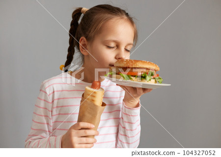 Charming cute beautiful little girl with pigtails with hot dog smelling aromat fresh baked hamburger standing with closed eyes isolated over gray background 104327052
