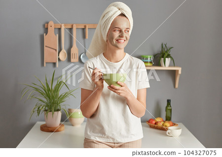 Smiling young woman posing in kitchen with towel on head having breakfast standing with plate in hands doing cosmetics procedures in the morning. 104327075
