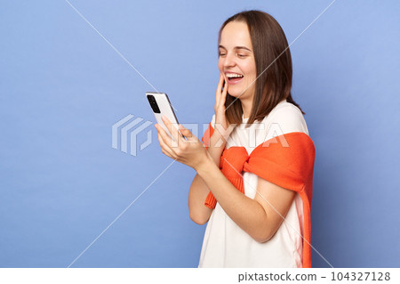 Laughing excited woman wearing white t-shirt and jumper over neck standing isolated over blue background using mobile phone checking social networks. Laughing excited woman wearing white t-shirt and jumper over neck standing isolated over blue background using mobile phone checking social networks. 104327128