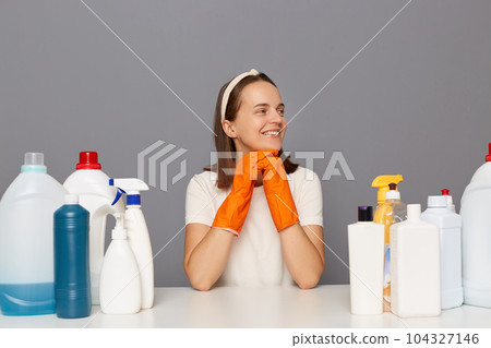 Smiling dreaming woman posing at workplace with cleaning detergents isolated over gray background finishing her household chores looking away with happy facial expression. Smiling dreaming woman posing at workplace with cleaning detergents isolated over gray background finishing her household chores looking away with happy facial expression. 104327146