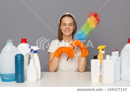 Joyful happy smiling woman posing at workplace with cleaning detergents sitting with pp duster in hands cleaning her house isolated over gray background. Joyful happy smiling woman posing at workplace with cleaning detergents sitting with pp duster in hands cleaning her house isolated over gray background. 104327147