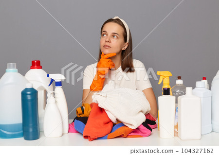 Pensive thoughtful woman posing at workplace with cleaning detergents isolated over gray background holding chin looking away thinking about household tasks. 104327162