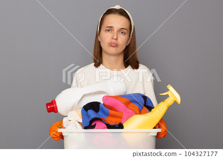 Exhausted woman wearing whote t-shirt with laundry bowl and detergent isolated over gray background feeling bored and unhappy needs rest looking at camera with sad expression. Exhausted woman wearing whote t-shirt with laundry bowl and detergent isolated over gray background feeling bored and unhappy needs rest looking at camera with sad expression. 104327177