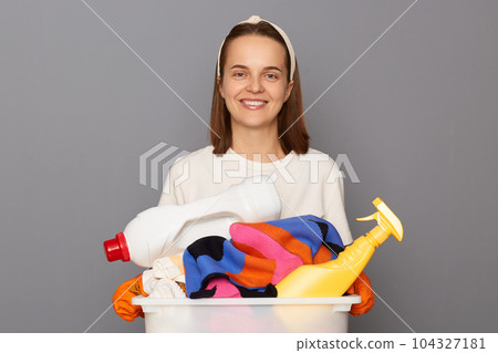 Routine washing keeps garments fresh. Housekeeper maintains clean and tidy house. Smiling cheerful woman wearing whote t-shirt with laundry bowl and detergent isolated over gray background Routine washing keeps garments fresh. Housekeeper maintains clean and tidy house. Smiling cheerful woman wearing whote t-shirt with laundry bowl and detergent isolated over gray background 104327181