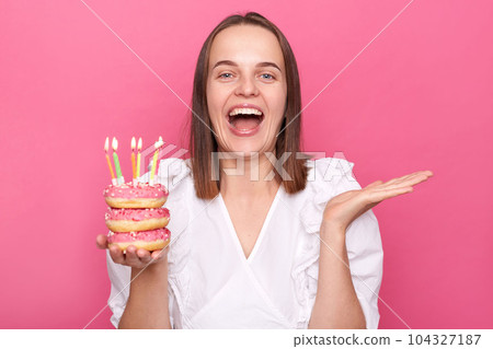 Satisfied brown haired woman holding delicious appetizing doughnuts with burning candles wears white blouse celebrating birthday alone at home isolated over pink background. 104327187