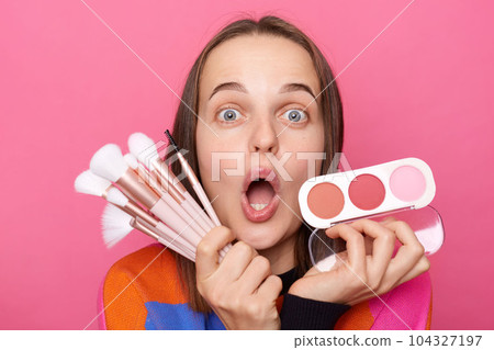 Shocked surprised woman wearing sweater holding brushes and powder posing isolated on pink background looking with big eyes, being amazed with new cosmetic products. 104327197
