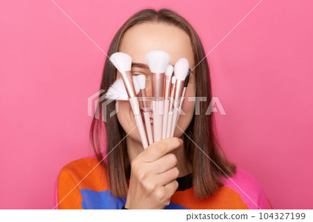 Positive woman wearing sweater hiding her face with cosmetic brushes posing isolated on pink background preparing for doing makeup, showing new tools for cosmetic procedures. 104327199