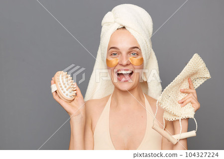 Excited joyful young woman wrapped head in towel with patches under eyes posing isolated over gray background, holding dry brush and shower sponge, doing spa procedures. Excited joyful young woman wrapped head in towel with patches under eyes posing isolated over gray background, holding dry brush and shower sponge, doing spa procedures. 104327224