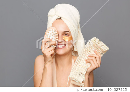 Spa treatment. Body care. Playful smiling young woman wrapped head in towel with patches under eyes covering eye with dry brush having in bathroom posing isolated over gray background. Spa treatment. Body care. Playful smiling young woman wrapped head in towel with patches under eyes covering eye with dry brush having in bathroom posing isolated over gray background. 104327225
