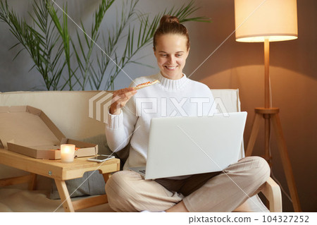 Relaxed happy smiling woman with bun hairstyle in white sweater looking at laptop screen and having snack eating pizza while watching film sitting on sofa at home. 104327252
