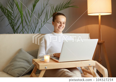 Smiling freelancer woman with bun hairstyle in white sweater looking at laptop screen enjoying her work communicating online sitting on sofa at home. Smiling freelancer woman with bun hairstyle in white sweater looking at laptop screen enjoying her work communicating online sitting on sofa at home. 104327254