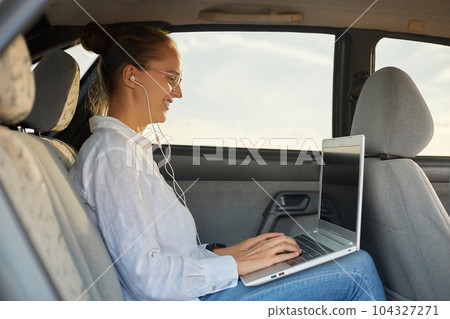 Side view portrait of confident woman working on laptop while sitting in the car listening music while riding to work smiling typing on keyboard freelance job. 104327271