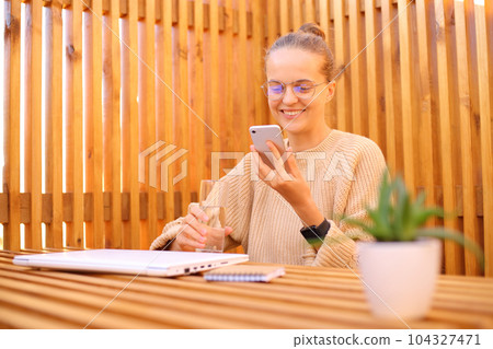 Good looking winsome beautiful woman wearing beige sweater sitting in outdoor cafe working on laptop and using mobile phone watching video checking social networks. 104327471