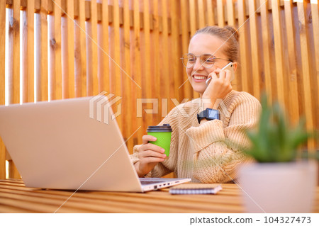 Attractive young woman wearing beige sweater sitting in outdoor cafe working on laptop, drinking coffee talking on mobile phone enjoying her freelance work having break. 104327473