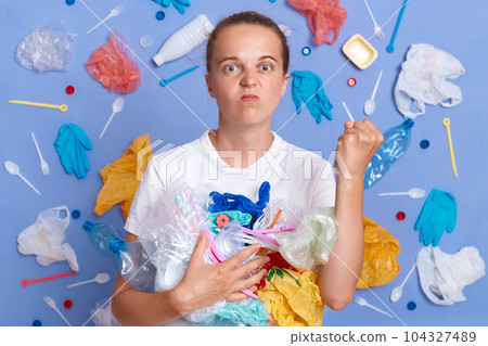 Volunteers clean up garbage. Reuse materials to reduce waste. Angry woman wearing white shirt isolated on blue wall with litter around with rubbish showing raised fist. 104327489