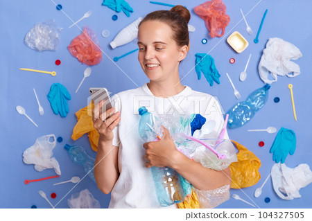 Positive smiling woman wearing white shirt isolated on blue wall with garbage around using mobile phone, cleaning up parks and gardens, saving planet. Positive smiling woman wearing white shirt isolated on blue wall with garbage around using mobile phone, cleaning up parks and gardens, saving planet. 104327505