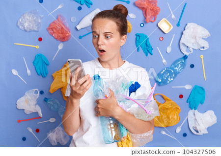 Reduce garbage pollution. Cleaning up planet. Shocked surprised woman wearing white shirt isolated on blue wall with garbage around looking at cell phone screen with open mouth. 104327506